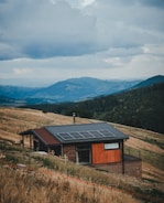 A small wooden house with a modern design is situated on a sloping grassy hillside. The house has solar panels installed on the roof. The landscape includes rolling hills and mountains in the distance, covered with dense forests. The sky is cloudy and overcast, suggesting an impending storm or recent rainfall.