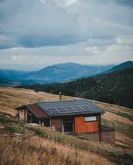 A small wooden house with a modern design is situated on a sloping grassy hillside. The house has solar panels installed on the roof. The landscape includes rolling hills and mountains in the distance, covered with dense forests. The sky is cloudy and overcast, suggesting an impending storm or recent rainfall.