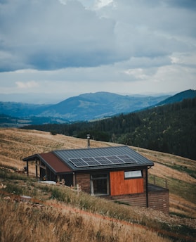 A small wooden house with a modern design is situated on a sloping grassy hillside. The house has solar panels installed on the roof. The landscape includes rolling hills and mountains in the distance, covered with dense forests. The sky is cloudy and overcast, suggesting an impending storm or recent rainfall.