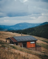A small wooden house with a modern design is situated on a sloping grassy hillside. The house has solar panels installed on the roof. The landscape includes rolling hills and mountains in the distance, covered with dense forests. The sky is cloudy and overcast, suggesting an impending storm or recent rainfall.