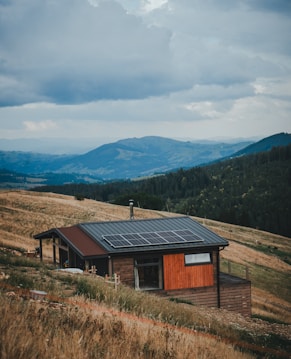 A small wooden house with a modern design is situated on a sloping grassy hillside. The house has solar panels installed on the roof. The landscape includes rolling hills and mountains in the distance, covered with dense forests. The sky is cloudy and overcast, suggesting an impending storm or recent rainfall.