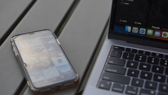 A smartphone with an app screen is placed next to a laptop with a visible keyboard and part of the screen. The phone displays various app icons and images, while the laptop shows a desktop with multiple application icons.