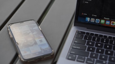 A smartphone with an app screen is placed next to a laptop with a visible keyboard and part of the screen. The phone displays various app icons and images, while the laptop shows a desktop with multiple application icons.