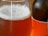 A close-up view of a glass filled with a frothy, amber-colored liquid, likely beer, with bubbles on the surface. A bottle with an orange label is partially visible in the background.