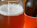 A close-up view of a glass filled with a frothy, amber-colored liquid, likely beer, with bubbles on the surface. A bottle with an orange label is partially visible in the background.