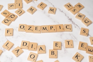 Wooden Scrabble tiles arranged on a white marble surface with the word 'OZEMPIC' in the center. Other scattered tiles spell out parts of words like 'DIABETES' and 'FOLD'. The arrangement of letters suggests a theme related to word games or puzzles.