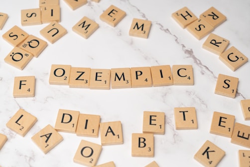 Wooden Scrabble tiles arranged on a white marble surface with the word 'OZEMPIC' in the center. Other scattered tiles spell out parts of words like 'DIABETES' and 'FOLD'. The arrangement of letters suggests a theme related to word games or puzzles.