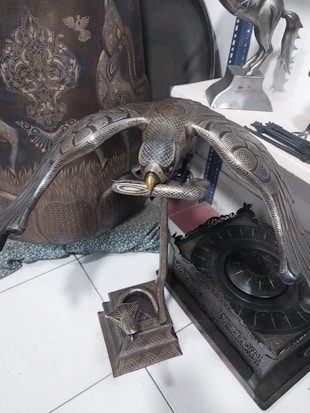 A detailed metallic sculpture of a bird with outstretched wings, crafted with intricate patterns and a lifelike pose. The sculpture is displayed indoors, surrounded by other ornate metalwork pieces, including a figurine of a horse and decorated plates.