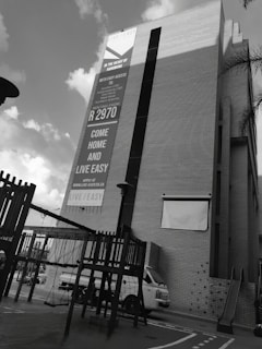 A tall building features a large advertisement promoting rental opportunities. The lower part of the scene includes playground equipment and a parked white van. The sky above is partly cloudy, creating a contrast with the building.