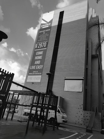 A tall building features a large advertisement promoting rental opportunities. The lower part of the scene includes playground equipment and a parked white van. The sky above is partly cloudy, creating a contrast with the building.
