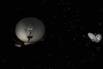 Two spacecraft with large dish antennas are in deep space surrounded by numerous small, bright stars against a dark background. The spacecraft appear to be similar in design, featuring round dishes and various scientific instruments attached to the bodies.