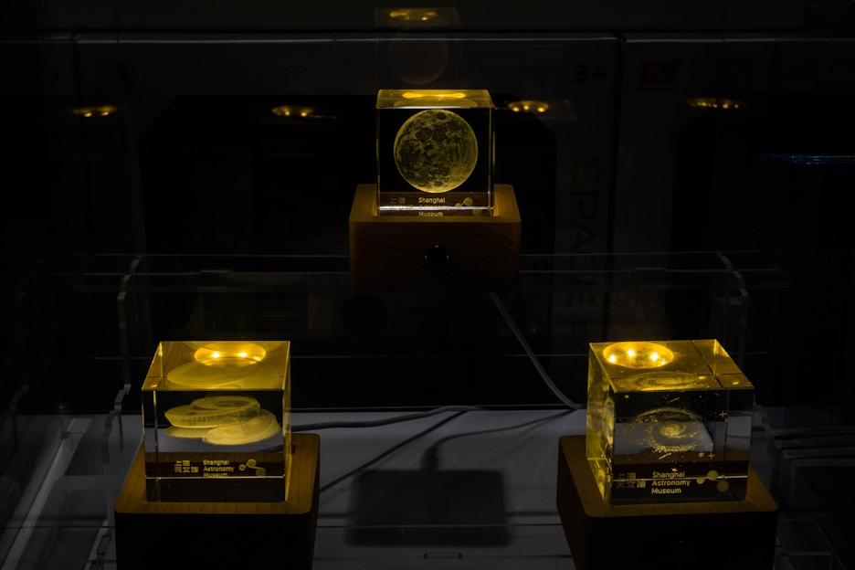 Three illuminated crystal cubes rest on wooden bases, each displaying intricate celestial designs that appear to be etched or embedded within the glass. The cubes are lit with a warm, yellow glow, emphasizing the details of the celestial objects, which resemble planets or moons. The background is dark, enhancing the visibility of the glowing cubes.
