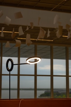Papers are suspended in mid-air, seemingly floating, above a modern-looking table lamp with a circular light source. The background shows large windows with a view of an overcast sky and a distant landscape.