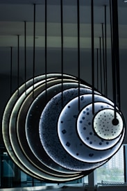 A modern art installation comprised of circular panels suspended from the ceiling. Each panel features different patterns and textures, resembling celestial bodies or abstract representations of planets. The installation uses gradients of dark and light colors, with the use of illumination enhancing its futuristic appearance. The setting appears to be indoors, possibly within a gallery or public space.