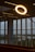 A glowing circular light fixture is prominently displayed indoors against the backdrop of large windows. The view through the windows includes a landscape with trees and a body of water. Blue lettering is visible on the window glass.