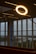 A glowing circular light fixture is prominently displayed indoors against the backdrop of large windows. The view through the windows includes a landscape with trees and a body of water. Blue lettering is visible on the window glass.