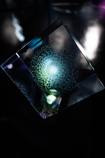 A geometric glass prism with a glowing blue center surrounded by a pattern of scattered speckles, set against a dark, reflective background.