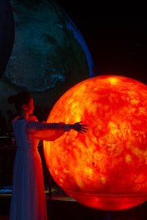 A person in a flowing white dress stands with extended arms toward a large, glowing orange-red sphere resembling the sun. The background shows another large earth-like sphere with a dark ambiance.