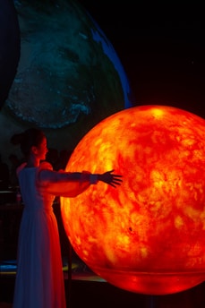 A person in a flowing white dress stands with extended arms toward a large, glowing orange-red sphere resembling the sun. The background shows another large earth-like sphere with a dark ambiance.