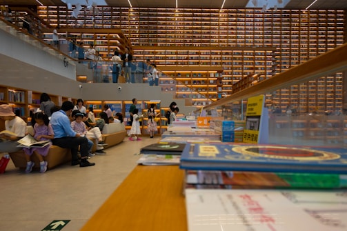 A large, modern library interior with high ceilings and an extensive wall of bookshelves. Several people are sitting and standing, some reading books or interacting with each other. The lighting is warm and inviting, with books and seating areas in the foreground and background.