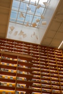 A library with towering wooden bookshelves filled with books and illuminated by warm lights. Above, a glass ceiling lets in natural light and features artistic paper-like sculptures suspended in motion, creating a sense of whimsical movement.