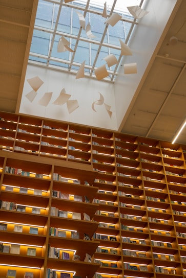 A library with towering wooden bookshelves filled with books and illuminated by warm lights. Above, a glass ceiling lets in natural light and features artistic paper-like sculptures suspended in motion, creating a sense of whimsical movement.
