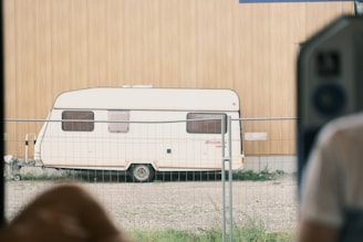 A white caravan is parked on a gravel surface, surrounded by a metal fence. The wall behind the caravan is made of light brown wood. A blurred portion of a person wearing a white shirt is visible on the right side, suggesting the photo is taken from inside a building or vehicle.