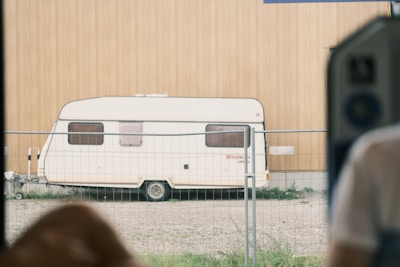 A white caravan is parked on a gravel surface, surrounded by a metal fence. The wall behind the caravan is made of light brown wood. A blurred portion of a person wearing a white shirt is visible on the right side, suggesting the photo is taken from inside a building or vehicle.