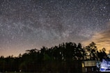 A star-filled sky spans across the night, with the faint outline of the Milky Way visible. Below, a silhouette of a dense forest creates a dark horizon against the brightly lit sky. To the right, a small structure with illuminated windows stands near the trees, and a gentle glow on the horizon suggests distant light pollution or a setting sun.