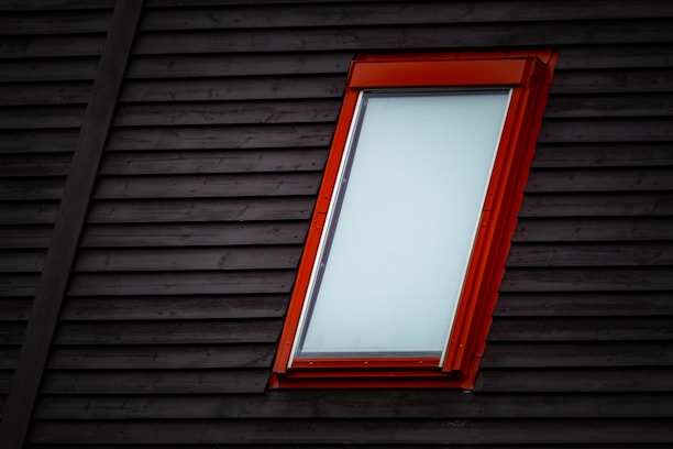 A modern window with a bright red frame is set into a dark wooden wall. The window is rectangular and mostly covered, reflecting a minimalistic architectural style.