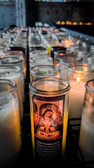 A collection of lit devotional candles arranged in rows inside a dimly lit space. One candle in particular is highlighted with an image of the Virgin Mary and Spanish text on its glass. The background features soft, warm light from the candles casting a gentle glow.