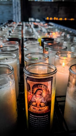 A collection of lit devotional candles arranged in rows inside a dimly lit space. One candle in particular is highlighted with an image of the Virgin Mary and Spanish text on its glass. The background features soft, warm light from the candles casting a gentle glow.