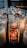 A collection of lit devotional candles arranged in rows inside a dimly lit space. One candle in particular is highlighted with an image of the Virgin Mary and Spanish text on its glass. The background features soft, warm light from the candles casting a gentle glow.