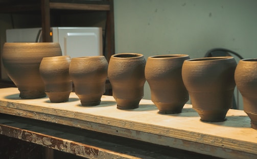 A row of handcrafted clay pots is displayed on a wooden shelf. The pots vary slightly in size and shape, each with a smooth surface and earthy matte finish. The background shows a cluttered workshop environment, suggesting a creative space dedicated to pottery.