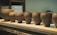 A row of handcrafted clay pots is displayed on a wooden shelf. The pots vary slightly in size and shape, each with a smooth surface and earthy matte finish. The background shows a cluttered workshop environment, suggesting a creative space dedicated to pottery.