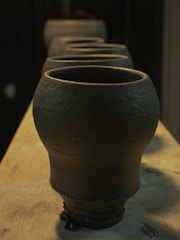 A series of clay pots are lined up on a wooden surface, displaying a rustic, textured finish. The lighting is warm, casting gentle shadows that accentuate the curves and rough surfaces of the pottery.