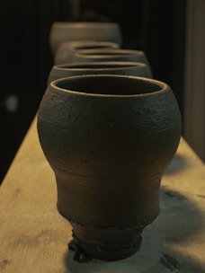 A series of clay pots are lined up on a wooden surface, displaying a rustic, textured finish. The lighting is warm, casting gentle shadows that accentuate the curves and rough surfaces of the pottery.