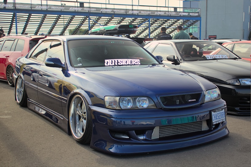A sleek blue car with polished wheels and lowered suspension is parked in an automotive event alongside other modified cars. The cars have decals and stickers displayed prominently on the windshields, and a small group of people can be seen in the background.