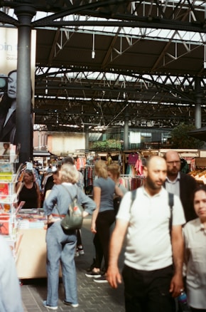 A busy indoor market scene with people walking and browsing stalls. Various items, including books and clothing, are displayed for sale. The market is housed in a space with a high, metal-truss roof.
