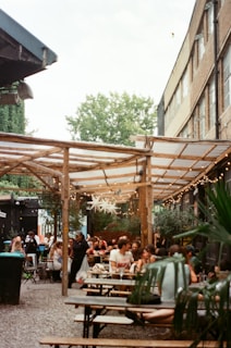 An outdoor courtyard with wooden benches and tables filled with people. The space is covered by a wooden pergola decorated with hanging string lights and star-shaped ornaments. Surrounding greenery includes trees and potted plants. The atmosphere appears casual and social.