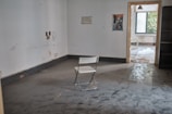 A sparsely furnished room with worn floors and an empty folding chair. The walls are bare except for a few marks and a small poster. There are openings leading to other areas of the building with visible greenery outside through the window.
