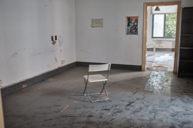 A sparsely furnished room with worn floors and an empty folding chair. The walls are bare except for a few marks and a small poster. There are openings leading to other areas of the building with visible greenery outside through the window.