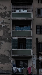 An aged residential building exterior featuring balconies with peeling paint and visible wear. Clothes hang on a line from the bottom balcony. The overall structure appears neglected, adding to an urban decay ambiance.
