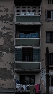 An aged residential building exterior featuring balconies with peeling paint and visible wear. Clothes hang on a line from the bottom balcony. The overall structure appears neglected, adding to an urban decay ambiance.