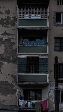 An aged residential building exterior featuring balconies with peeling paint and visible wear. Clothes hang on a line from the bottom balcony. The overall structure appears neglected, adding to an urban decay ambiance.
