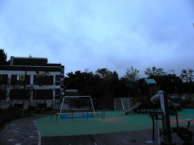 A playground with various play structures is situated next to a building. The structures include a slide, swings, and climbing equipment. The ground is covered with green and orange safety flooring, and there is a path made of paving stones. The background features trees and a cloudy sky, suggesting overcast weather.
