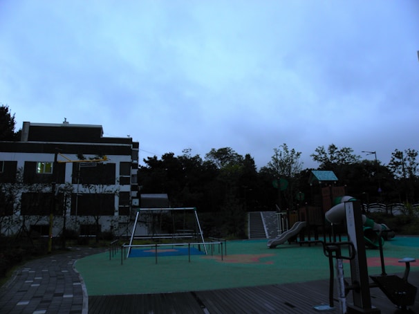 A playground with various play structures is situated next to a building. The structures include a slide, swings, and climbing equipment. The ground is covered with green and orange safety flooring, and there is a path made of paving stones. The background features trees and a cloudy sky, suggesting overcast weather.