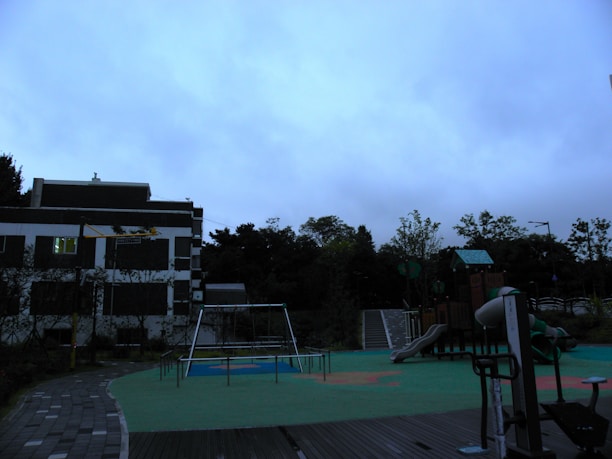 A playground with various play structures is situated next to a building. The structures include a slide, swings, and climbing equipment. The ground is covered with green and orange safety flooring, and there is a path made of paving stones. The background features trees and a cloudy sky, suggesting overcast weather.