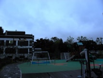A playground with various play structures is situated next to a building. The structures include a slide, swings, and climbing equipment. The ground is covered with green and orange safety flooring, and there is a path made of paving stones. The background features trees and a cloudy sky, suggesting overcast weather.