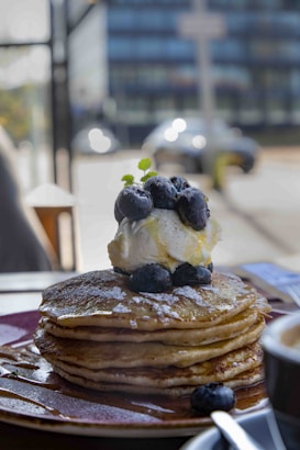 A stack of pancakes topped with fresh blueberries and a dollop of whipped cream, garnished with a small leaf. The pancakes are drizzled with syrup, and a cup of coffee is visible nearby. The setting appears to be a café with an urban background visible through a window.